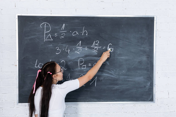A teenage girl is solving a math problem at school by writing on a school blackboard with white chalk. Her hair is pinned with a pink ribbon in two ponytails. - Фото, изображение