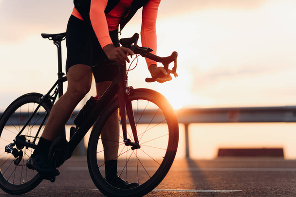Primo piano di atleta maschile in abiti da ciclismo luminosi e scarpe da ginnastica da corsa su strada con sorprendente tramonto sullo sfondo. Concetto di hobby preferito. - Foto, immagini