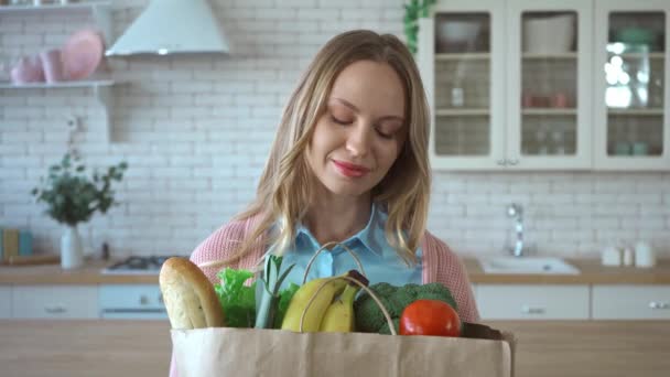 Donna sorridente con generi alimentari in shopping bag guardando la fotocamera in cucina - Filmati, video