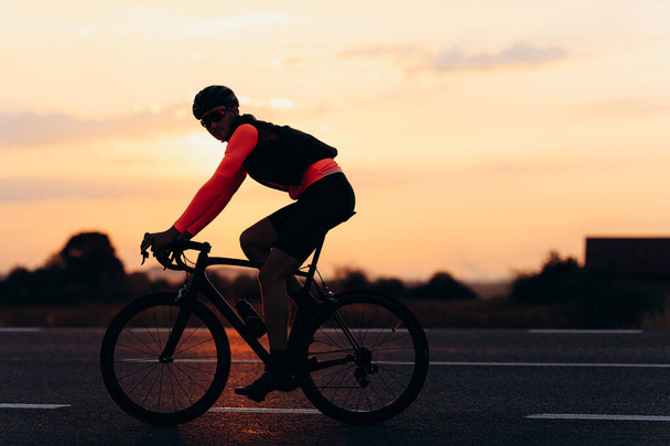 Bello il giovane ciclista che indossa casco protettivo e occhiali da ciclismo su strada all'aperto. Sfondo di bel tramonto. Concetto di corsa. - Foto, immagini