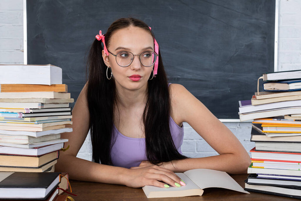 A teenager sits in the classroom among the piles of books and begins reading the first reading. The student starts learning. Her hair is pinned with a pink ribbon in two ponytails. - Foto, imagen
