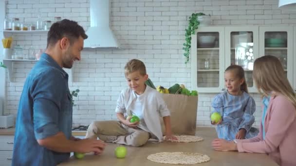 Feliz padre haciendo malabares con manzanas verdes cerca de la familia emocionada en la cocina - Imágenes, Vídeo
