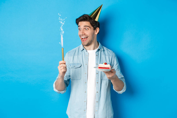 Glücklicher junger Mann feiert Geburtstag in Partyhut, hält B-Day-Kuchen und lächelt, steht über blauem Hintergrund - Foto, Bild