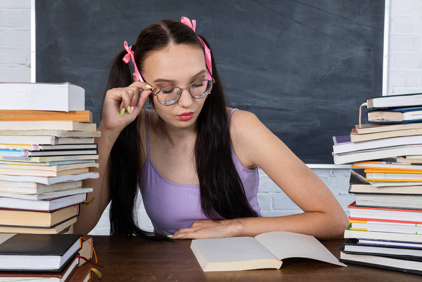 A teenager sits in the classroom among the piles of books and begins reading the first reading. The student starts learning. Her hair is pinned with a pink ribbon in two ponytails. - Фото, изображение