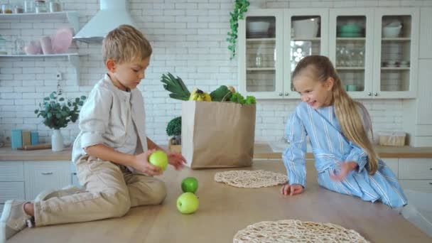 Χαρούμενα αδέλφια ρίχνουν πράσινα μήλα στο τραπέζι κοντά τσάντα ψώνια στην κουζίνα - Πλάνα, βίντεο
