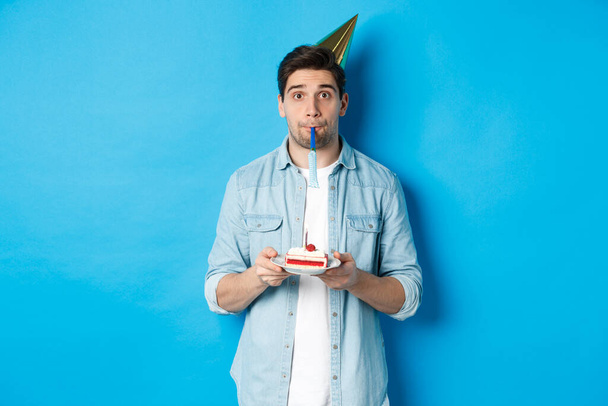 Bello giovane che tiene la torta di compleanno, indossa il cappello da festa e fischia, in piedi sopra lo sfondo blu - Foto, immagini