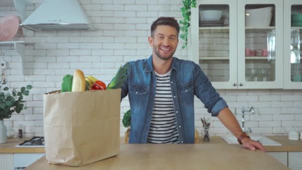 Veselý muž při pohledu na kameru v blízkosti nákupní tašky s potravinami v kuchyni - Záběry, video