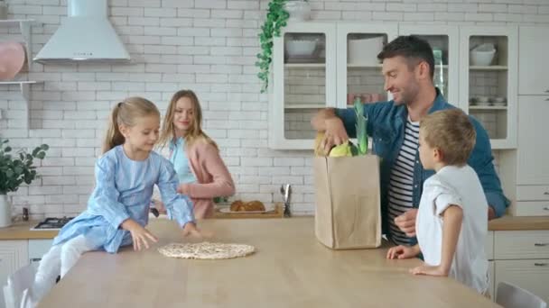 Mosolygó apa kicsomagolás bevásárló táska élelmiszerekkel az asztalon a család közelében - Felvétel, videó