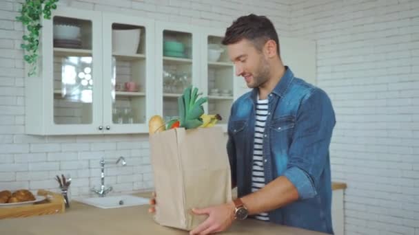 Uomo sorridente disimballaggio shopping bag con generi alimentari in cucina - Filmati, video