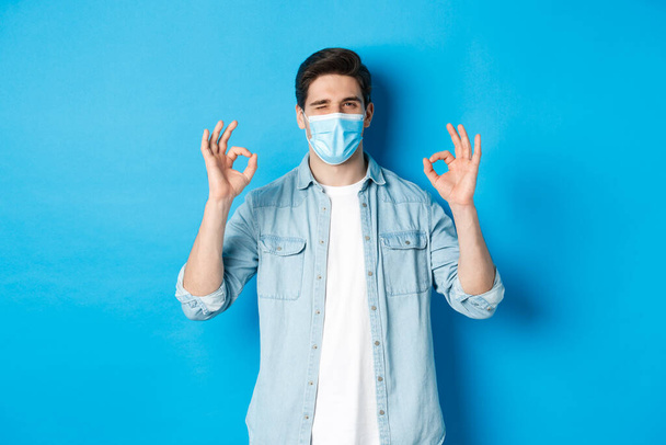 Concept of coronavirus, quarantine and social distancing. Cheeky man in medical mask winking, showing okay signs, assure or guarantee something, like and approve - 写真・画像