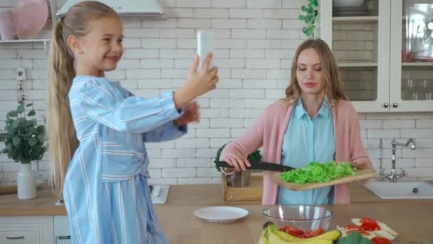 Ευτυχισμένη κόρη με smartphone λήψη φωτογραφιών της μητέρας προετοιμασία σαλάτα στην κουζίνα - Πλάνα, βίντεο