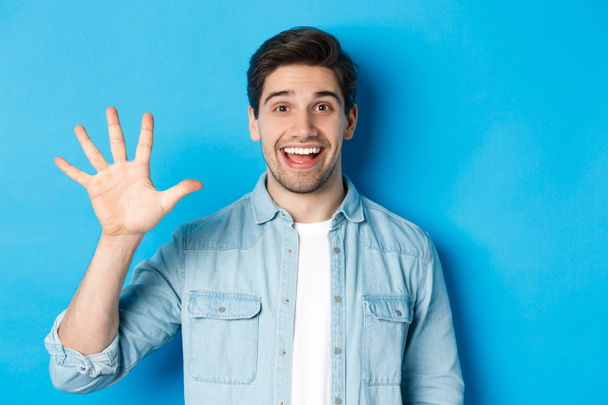 Közelkép jóképű férfi mosolyog, mutatja ujjak száma 5, álló felett kék háttér - Fotó, kép