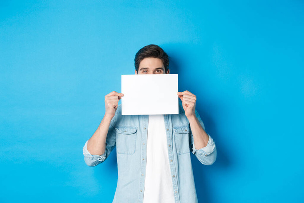 Чихлый красивый парень прячет лицо за пустым листом бумаги для вашего логотипа, делает объявление или показывает промо-предложение, стоя на синем фоне - Фото, изображение