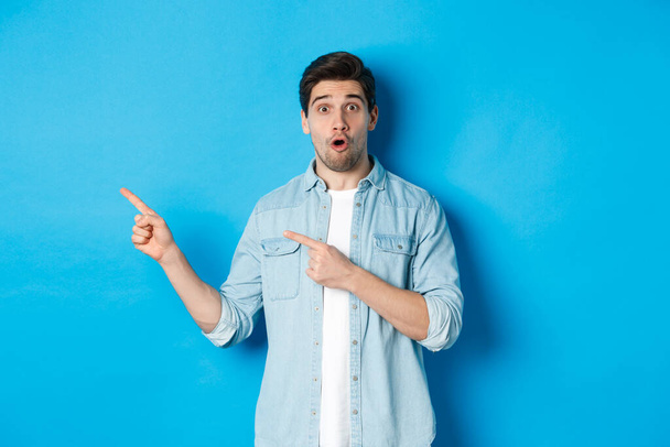 Portrait of surprised adult man in casual clothes showing announcement, pointing fingers left and looking amazed, standing against blue background - Photo, Image