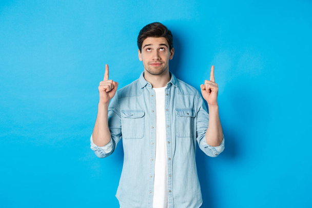 Portrait of displeased and skeptical male model pointing fingers up, looking at something unpleasant, standing against blue background - Foto, afbeelding