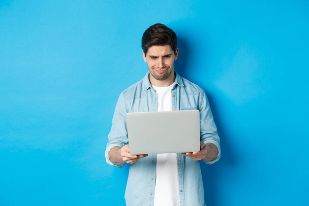 Απογοητευμένος όμορφος άντρας κοιτάζει την οθόνη του φορητού υπολογιστή και γκρινιάζει, κρίνοντας κάτι κακό στο διαδίκτυο, στέκεται πάνω από το μπλε φόντο - Φωτογραφία, εικόνα