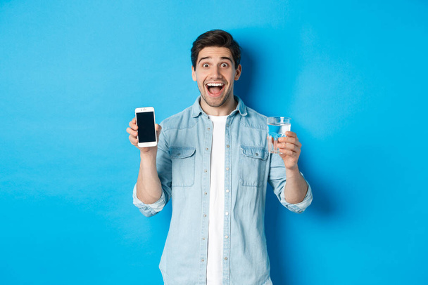 水とモバイル画面のガラスを示す幸せな男,スマートフォンの健康アプリをお勧めします,青の背景の上に立って - 写真・画像