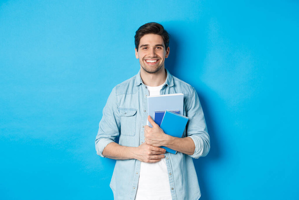 Молодой человек, держащий тетради и учебные материалы, улыбающийся счастливый, стоящий на синем фоне - Фото, изображение