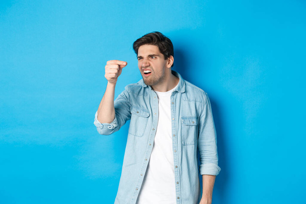 Confused guy holding something tiny in fingers and looking at it puzzled, standing over blue background - Photo, Image