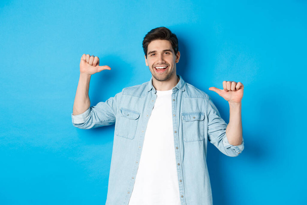 Portrait of confident man pointing at himself, being a professional, bragging while standing against blue background - Фото, изображение