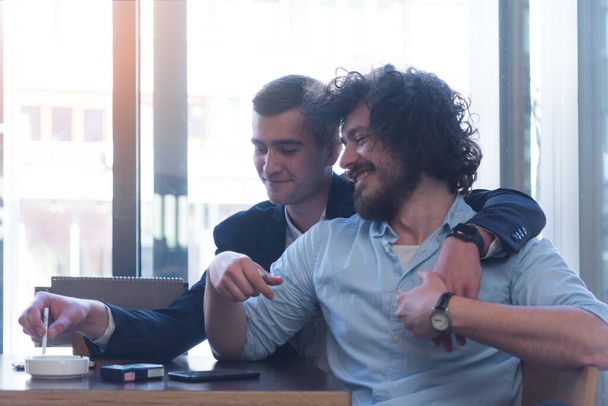 Δύο γκέι άντρες φίλοι αγκαλιασμένοι σε ένα καφέ, χαμογελώντας και κάνοντας ένα τσιγάρο. ΛΟΑΤ και έννοια της αγάπης. - Φωτογραφία, εικόνα