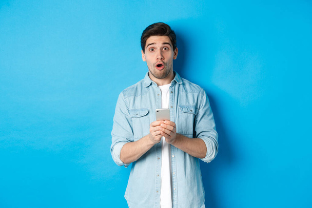 Verrast man in casual kleding kijken verbaasd, met smartphone, staande tegen een blauwe achtergrond - Foto, afbeelding
