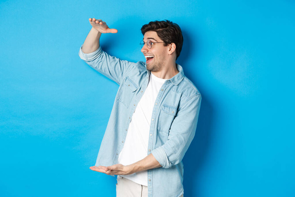Excited handsome man showing big size object and looking amazed, standing over blue background - Photo, Image