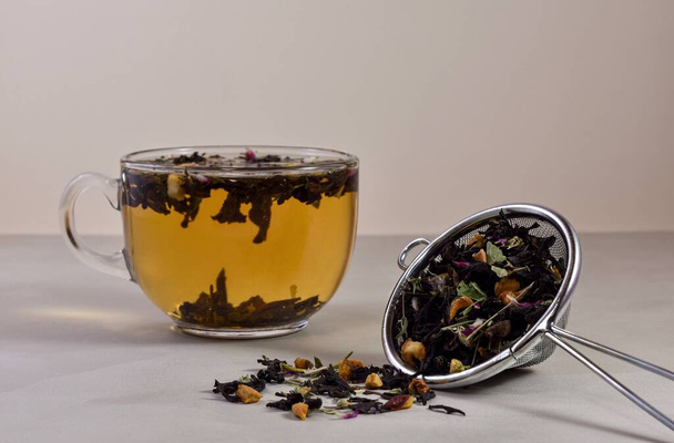 A glass, transparent cup with hot brewed tea, a metal strainer with a dry mix of herbs, fruits and flowers on a light beige background. Tea ceremony concept. - Foto, afbeelding