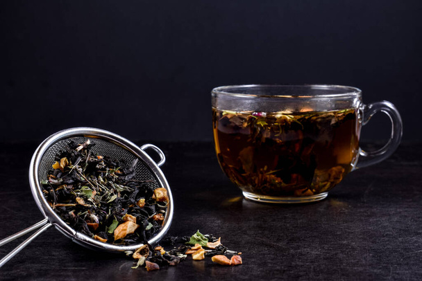 Glass cup with hot brewed tea, metal strainer with dry mix of herbs and fruits on a black wooden background. Concept for tea time. - Photo, image