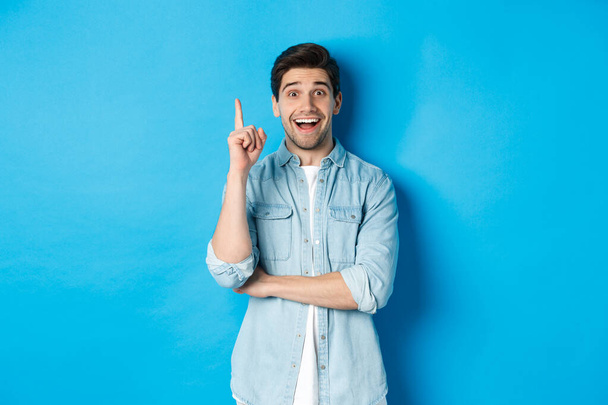 Image d'un bel homme adulte ayant une idée, levant le doigt et souriant excité, solution trouvée, debout sur fond bleu - Photo, image