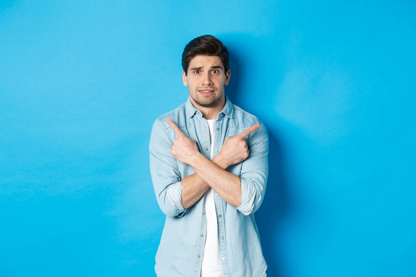 Portrait of nervous guy pointing fingers sideways, looking indecisive and asking for help with choice, showing left and right promo offers, standing against blue background - Foto, afbeelding