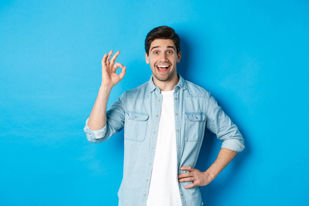 Glimlachende vrolijke man die ok teken toont en tevreden kijkt, iets goeds goedkeurt, tegen een blauwe achtergrond staat - Foto, afbeelding