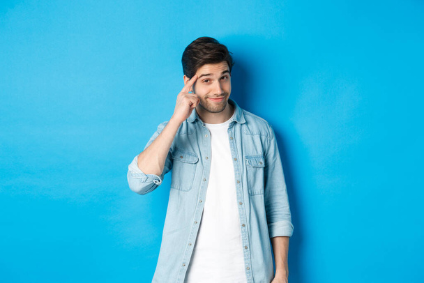 Young thoughtful man pointing at head, asking to think about it, giving a hint, standing in casual clothes over blue background - Photo, Image