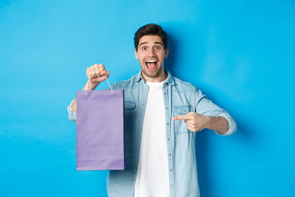 Concept of shopping, holidays and lifestyle. Excited guy pointing finger at paper bag and looking amazed, recommending store, announcing discounts, blue background - Zdjęcie, obraz