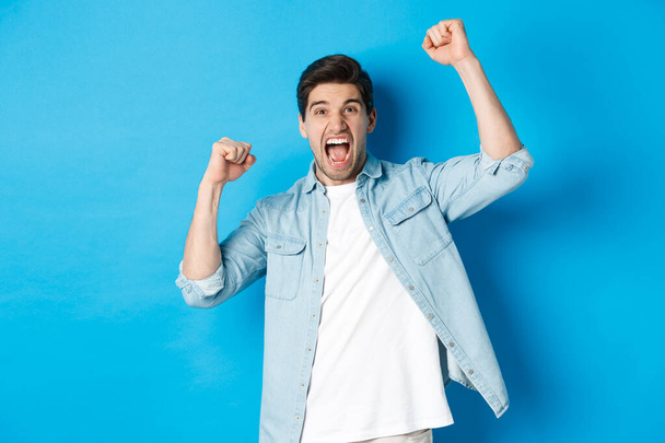 Cheerful guy making fist pumps and rooting for someone, shouting for joy, triumphing over win, standing against blue background - Foto, afbeelding