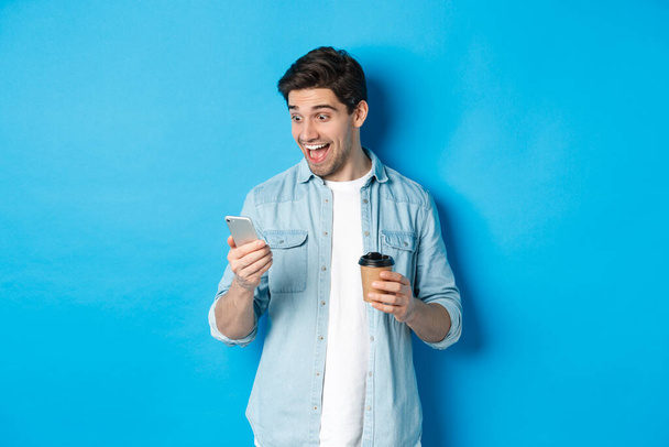 Mann blickt überrascht auf Smartphone, trinkt Kaffee, steht erstaunt vor blauem Hintergrund - Foto, Bild
