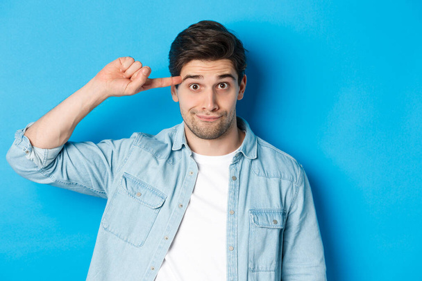 Close-up of man scolding for acting stupid or crazy, rolling finger on head and looking at camera, standing over blue background - Photo, Image