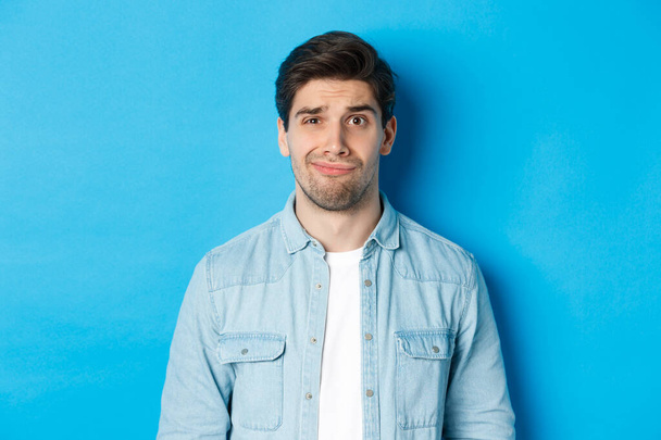 Close-up of skeptical and awkward guy smirking, feeling uncomfortable, standing over blue background - Foto, Imagem