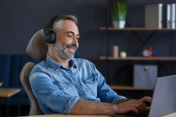 Portrait of handsome grey-haired man wearing headphones sitting at workplace. Senior male freelancer working in home office with laptop. Studying online, online courses - 写真・画像
