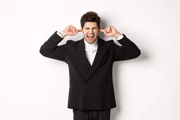Portret van geïrriteerde en lastige zakenman in zwart pak, oren dicht en schreeuwen, luid geklaag, staan tegen witte achtergrond - Foto, afbeelding