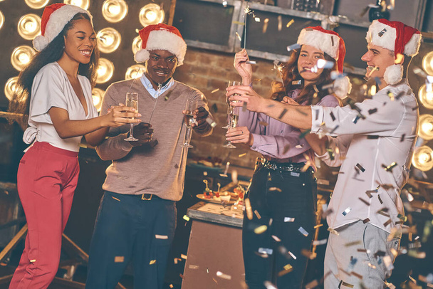 Happy New Year to you! Group of cheerful young people in santa hats carrying sparkling wine and bengal lights/firework. Party time. Celebrate concept - Fotoğraf, Görsel