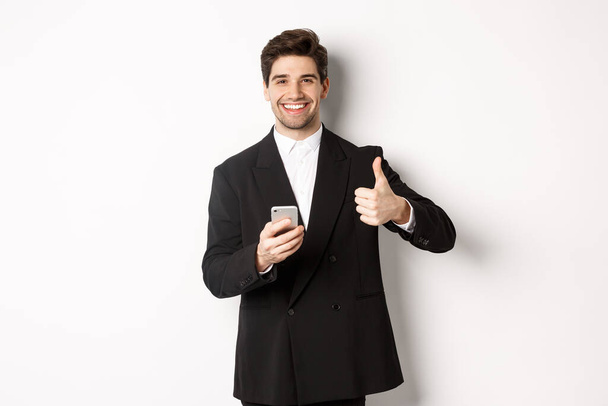 Schöner Typ im schicken Anzug, der Komplimente macht, Daumen hoch zeigt, während er sein Handy benutzt, eine App oder einen Online-Shop empfiehlt, vor weißem Hintergrund steht - Foto, Bild