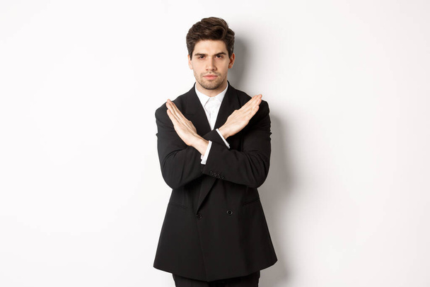 Portrait of serious and concerned man in black suit, showing stop gesture and frowning, making cross to prohibit or decline something, standing over white background - Foto, imagen