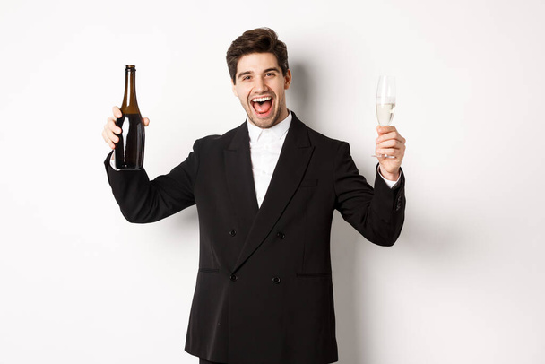 Concepto de vacaciones, fiesta y celebración. Hombre guapo en traje de moda que se divierte, sosteniendo la botella y la copa de champán, de pie sobre fondo blanco - Foto, imagen