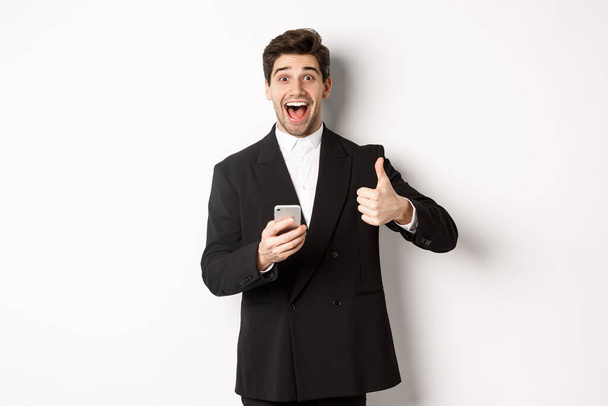 Retrato de hombre guapo en traje de moda que muestra los pulgares hacia arriba en la aprobación, utilizando la aplicación de teléfono móvil, sonriendo complacido, de pie sobre fondo blanco - Foto, imagen