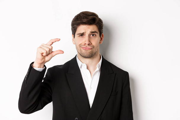 Close-up of handsome businessman in trendy suit, showing something small and looking disappointed, standing against white background - Foto, afbeelding