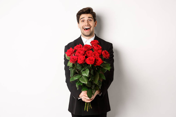 Image of handsome boyfriend in black suit, holding bouquet of red roses and smiling, being on a date, standing over white background - Photo, image