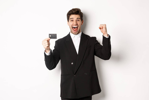 Portret van een opgewonden knappe zakenman in pak, blij en met een creditcard, staande tegen een witte achtergrond - Foto, afbeelding