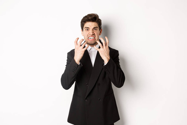 Image of angry businessman in suit, looking with furious expression and clenching fists, express hatred, standing mad over white background - Foto, immagini