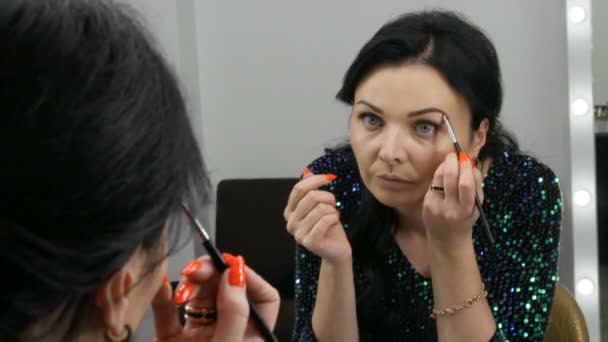 Joven hermosa mujer vistosa delante del espejo se aplica maquillaje de cara y ojos con un cepillo especial - Imágenes, Vídeo